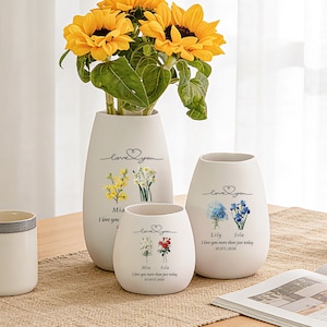 Op de afbeelding: Drie witte keramische vazen van verschillende hoogtes, elk voorzien van een "love you"-tekst, bloemenillustraties en gepersonaliseerde namen en data. De hoogste vaas bevat een boeket met felgele zonnebloemen en groene bladeren.