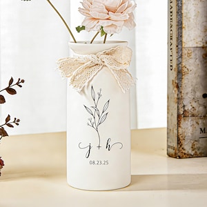 Könnte beinhalten: Eine zylindrische weiße Keramikvase mit einer schwarzen botanischen Strichzeichnung, den Kleinbuchstaben j + h und dem Datum 08.23.25 auf der Vorderseite. Ein beiges Spitzenband ist als Schleife um den Hals der Vase gebunden.