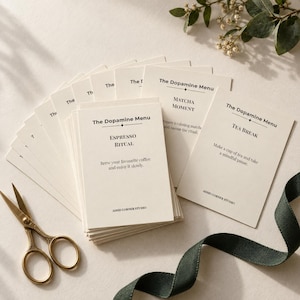 Könnte beinhalten: Ein Stapel cremefarbener, rechteckiger Karten mit der Überschrift The Dopamine Menu und schwarzem Text wie Espresso Ritual, Matcha Moment und Tea Break. Jede Karte enthält den Text Adhd Corner Studio. Eine goldene Schere liegt daneben.