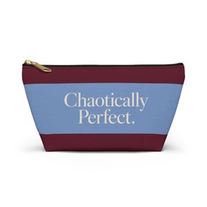 May include: A zippered pouch featuring a horizontal color-blocked design with a wide light blue center stripe flanked by dark maroon sections. The text "Chaotically Perfect." is printed in a cream-colored serif font across the light blue stripe.