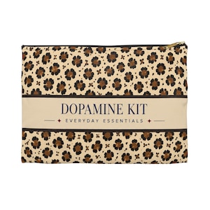 May include: A rectangular zippered pouch featuring a tan and brown leopard print pattern. A central horizontal band contains the dark blue text "DOPAMINE KIT" above the smaller text "EVERYDAY ESSENTIALS" flanked by two small four-pointed star icons.