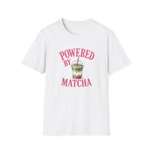 May include: A white short-sleeve t-shirt featuring a graphic of a layered matcha drink in a clear plastic cup with a pink straw. The text "POWERED BY MATCHA" is printed in pink capital letters above and below the drink illustration.