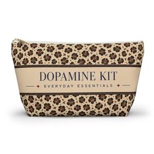 May include: A zippered pouch featuring a tan and brown leopard print pattern. A central horizontal band in a lighter tan shade displays the dark blue text DOPAMINE KIT and the text EVERYDAY ESSENTIALS, accented by two small four-pointed stars.
