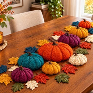 Pode incluir: Uma coleção de abóboras de tecido em várias cores, incluindo laranja, azul-petróleo, roxo, amarelo e creme, dispostas com folhas de outono de tecido sobre uma superfície de madeira. As abóboras possuem hastes castanhas.