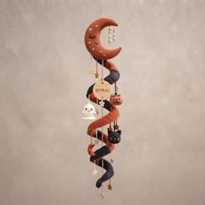 May include: A hanging crochet mobile featuring a large crescent moon at the top, followed by a long, spiraling structure in alternating rust orange and dark charcoal colors. Small crocheted figures of a ghost, pumpkin, and cat are attached.