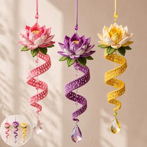 May include: Three hanging crochet lotus flower ornaments in pink, purple, and yellow. Each flower features green leaves, a spiraling crochet tail, and a clear teardrop-shaped crystal prism at the bottom, suspended by a matching colored string.