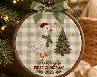 May include: A round wooden embroidery hoop ornament featuring a white and sage green gingham fabric background. It displays an embroidered white goose wearing a red Santa hat and green scarf, a decorated Christmas tree, and the text Avery's FIRST CHRISTMAS 2026, topped with a sage green satin ribbon bow.