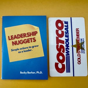 May include: A blue book titled Leadership Nuggets: Simple actions to grow as a leader by Becky Barker, Ph.D. next to a white Costco Wholesale Gold Star Member card. The book features a yellow sticky note graphic with red and blue text.