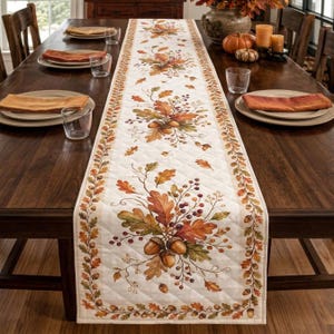 May include: A quilted cream-colored table runner featuring a repeating autumn-themed pattern of orange, brown, and yellow oak leaves, acorns, and berries. The runner has a decorative border of small leaves and acorns along its entire length.