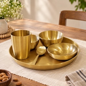 May include: A brass dinnerware set consisting of a large round tray, a tall tumbler, two small bowls, and a spoon. Each piece features a smooth, brushed metallic gold finish with a consistent, uniform appearance across the entire collection.