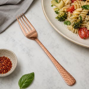 May include: A copper-toned metal fork with a long, slender handle featuring a textured, hammered finish. The fork has four tines and a polished, reflective surface that catches the light, positioned diagonally across a light-colored surface.