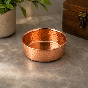 May include: A round, shallow bowl crafted from polished copper with a distinct hammered texture across its entire exterior and interior surface, featuring a smooth, flat base and a consistent metallic orange-brown finish throughout the vessel.