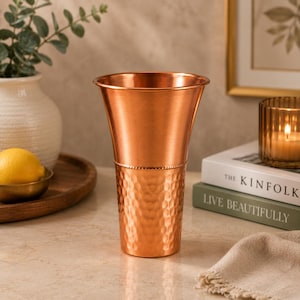 May include: A tall, conical copper vessel featuring a smooth upper section and a hammered, textured lower section separated by a thin beaded band. The metallic surface has a warm, reddish-brown finish that reflects light across its cylindrical form.