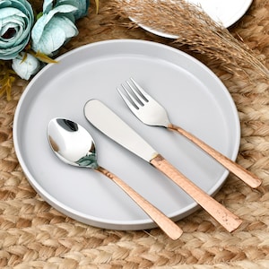 May include: A set of three stainless steel utensils including a spoon, a butter knife, and a fork. Each piece features a polished silver-toned head and a textured, copper-coloured metal handle, arranged neatly on a smooth, round, light grey plate.