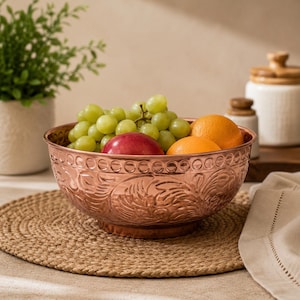 May include: A round copper bowl with an embossed floral pattern and a decorative circular border along the rim. The bowl is filled with a cluster of green grapes, a red apple, and two orange citrus fruits, showcasing a metallic finish.