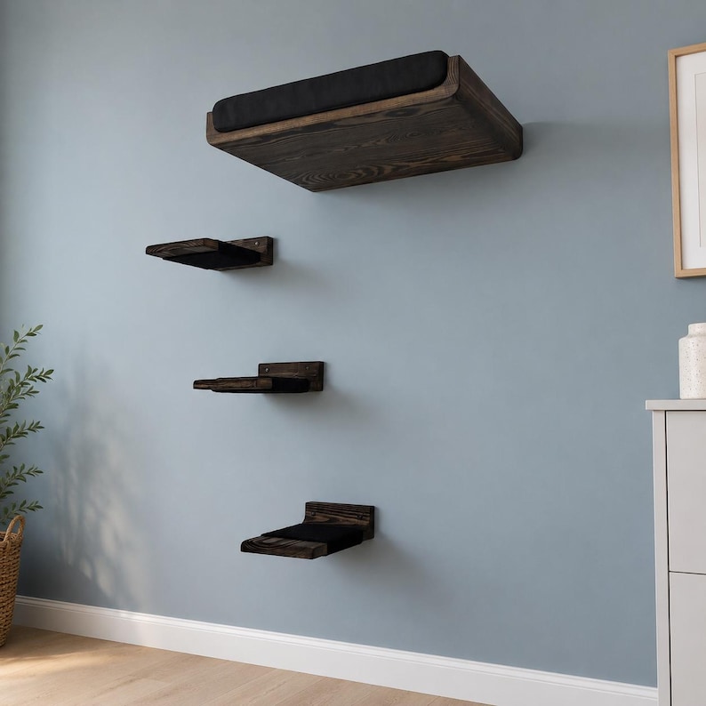 May include: A set of four wall-mounted floating shelves made of dark stained wood with visible grain. Each shelf features a rectangular black fabric cushion on the top surface, providing a soft platform for each of the four tiered wooden levels.