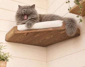 May include: A wall-mounted corner cat shelf made of dark stained wood with a visible grain pattern. The shelf features a soft, rectangular white cushion on top, providing a comfortable resting area for a fluffy grey cat currently yawning.
