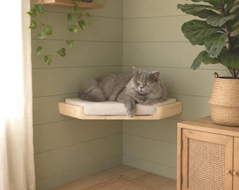 May include: A fluffy grey cat resting on a light-colored wooden corner shelf. The shelf features a soft, rectangular white cushion fitted to its surface. The cat is positioned in the center of the shelf, which is mounted against a sage green wall.