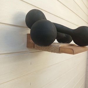 May include: A black dumbbell resting on a small, wall-mounted wooden shelf. The shelf features a light brown wood grain finish and a central section wrapped in thick, natural-colored jute rope, providing a textured surface for the weight.