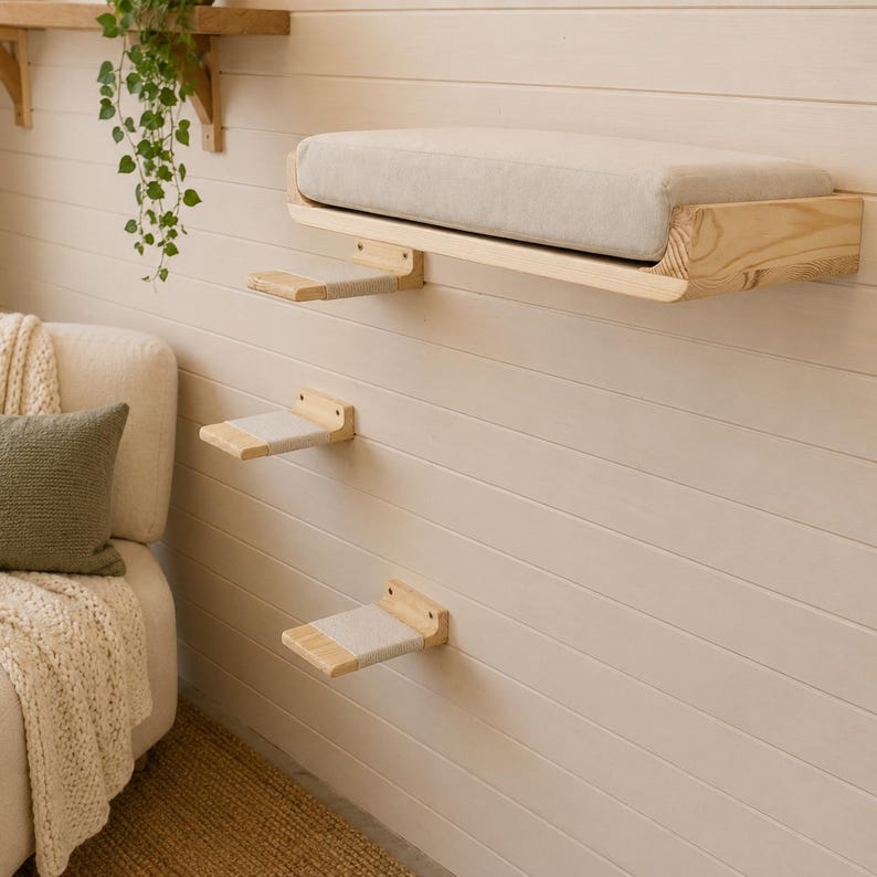 May include: A wall-mounted cat furniture set consisting of one large rectangular wooden shelf with a thick beige cushion and two smaller wooden steps, each featuring a beige fabric covering on the top surface for added grip and comfort.