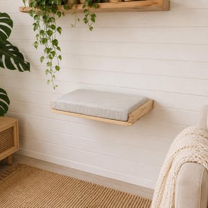 May include: A wall-mounted rectangular wooden shelf featuring a light gray fabric cushion on its top surface. The shelf is constructed from natural light-toned wood with a visible grain, providing a flat platform attached to a white paneled wall.