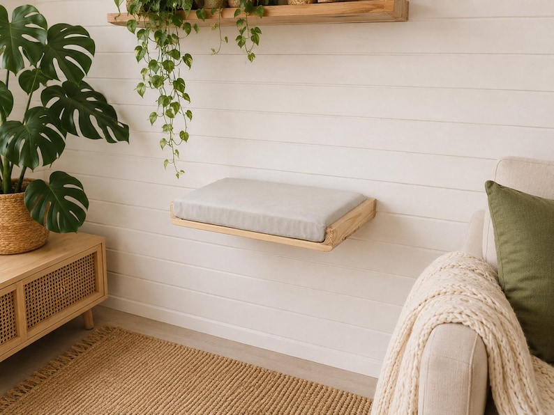 May include: A wall-mounted rectangular wooden shelf featuring a light gray fabric cushion on its top surface. The shelf is constructed from natural light-toned wood with a visible grain, providing a flat platform attached to a white paneled wall.