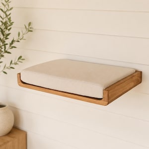 Può includere: Una mensola rettangolare da parete realizzata in legno naturale con finitura liscia. La mensola presenta un cuscino spesso rivestito in tessuto beige che si inserisce perfettamente nella cornice in legno, offrendo una superficie piana e imbottita.
