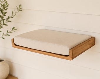 May include: A rectangular wall-mounted shelf made of natural wood with a smooth finish. The shelf features a thick, beige fabric-covered cushion that fits snugly within the wooden frame, providing a flat, padded surface for resting or display.