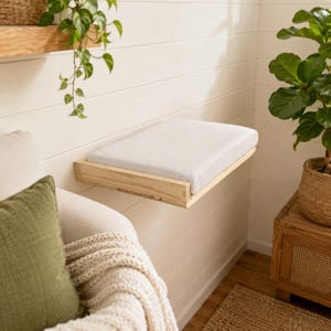 May include: A rectangular wall-mounted shelf featuring a light-colored wooden frame and a soft, light gray padded cushion top. The shelf is attached to a white horizontal paneled wall, providing a flat surface for resting items or pets.