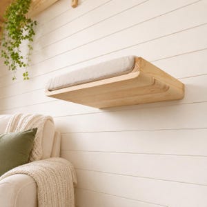 May include: A wall-mounted cat shelf made of light-colored natural wood with a rectangular shape. The shelf features a soft, beige fabric cushion securely placed on its top surface, providing a flat resting area for a pet to lounge upon.