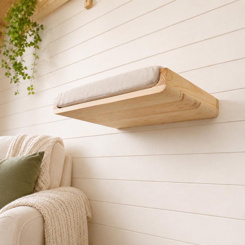 May include: A wall-mounted cat shelf made of light-colored natural wood with a rectangular shape. The shelf features a soft, beige fabric cushion securely placed on its top surface, providing a flat resting area for a pet to lounge upon.