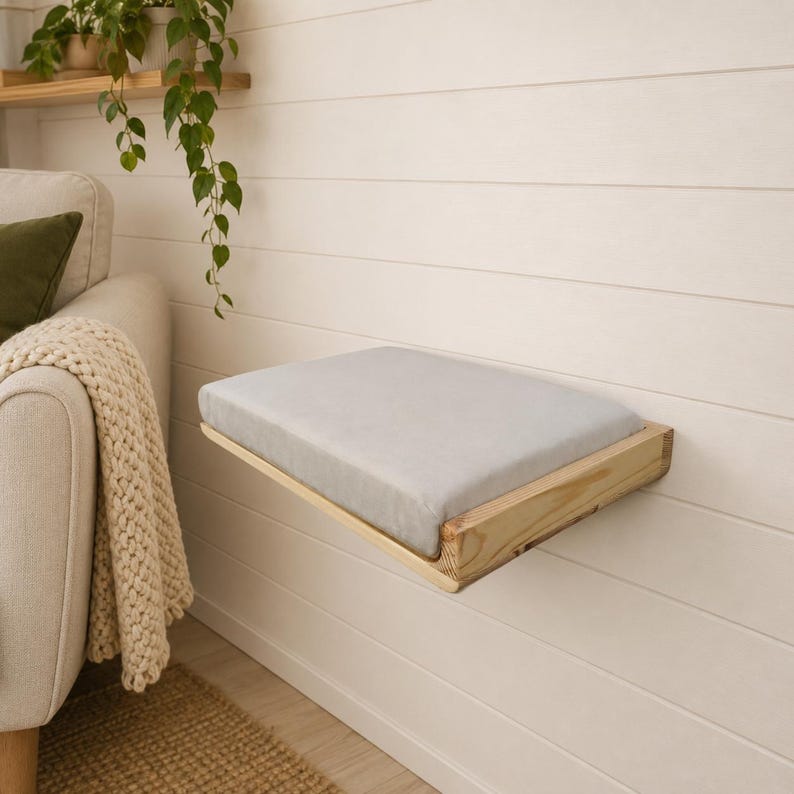 May include: A rectangular wall-mounted shelf featuring a light-colored wooden frame and a soft, light gray padded cushion top. The shelf is attached to a white horizontal paneled wall, providing a flat surface for resting or display purposes.