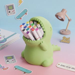 May include: A light green dinosaur-shaped desk organiser with an open mouth holding a collection of white markers with coloured caps. The organiser features a rounded body, small arms, and a tail, serving as a functional holder for art supplies.