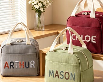 May include: Three rectangular lunch bags with cream-coloured handles and zippers. The grey bag features the name ARTHUR, the green bag features MASON, and the maroon bag features CHLOE in embroidered, multi-coloured, textured lettering.