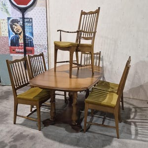 May include: A wooden dining set comprising an oval table with a central pedestal base and four chairs with spindle backs and olive green upholstered seats. One chair is placed on top of the table, while three chairs are arranged around it.