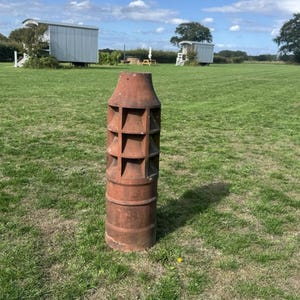 May include: A tall, cylindrical terracotta chimney pot with a weathered, reddish-brown finish. The upper section features a series of vertical and horizontal dividers forming a grid-like pattern, while the lower portion is a solid, smooth cylinder.