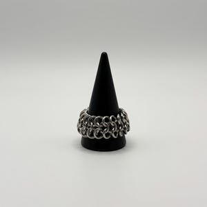 Könnte beinhalten: Ein silberfarbener Ring, der aus einer Reihe ineinandergreifender Metallringe besteht und ein kettenhemdartiges Muster bildet. Das Band weist ein gleichmäßiges, sich wiederholendes kreisförmiges Gliedermuster auf, das den gesamten Ring umschließt.