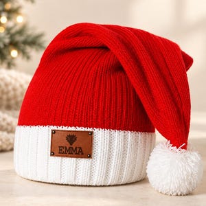 May include: A bright red and white ribbed knit beanie with a long, slouchy design and a white pom-pom at the tip. A rectangular brown leather patch is attached to the front white cuff, featuring a small graphic icon and the name EMMA in black.