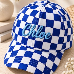 Puede incluir: Una gorra de béisbol con un patrón de cuadros azules y blancos en toda la superficie. El panel frontal muestra el nombre Chloe bordado en relieve con una fuente cursiva azul claro y un contorno azul más oscuro, centrado en la corona.