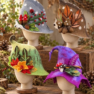 May include: Four felted hats displayed on mannequin heads. One is gray with a ladybug and red berries, one brown with autumn leaves and mushrooms, one green with orange flowers and leaves, and one purple with mushrooms and green foliage.