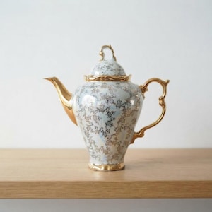 Op de afbeelding: Een porseleinen theepot met een witte basis en een ingewikkeld bloemmotief in goud en grijs. De theepot is voorzien van een bijpassend deksel met een gouden knop, een goudkleurige tuit en een gebogen gouden handvat, afgewerkt met glanzend glazuur.