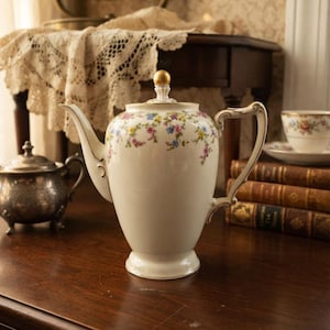 Może przedstawiać: Kremowy dzbanek do herbaty z porcelany z delikatnym wzorem kwiatowych girland w kolorach różowym, niebieskim i żółtym wokół górnej części. Dzbanek posiada złoty uchwyt na pokrywce, zakrzywioną rączkę oraz zwężający się dzióbek ze złotym wykończeniem.