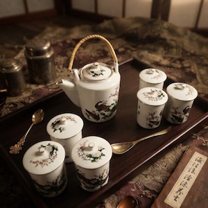 Peut inclure: Un service à thé en céramique blanche comprenant une théière avec une anse tressée et cinq tasses assorties avec couvercles, ornées de motifs floraux et d'oiseaux. L'ensemble est disposé sur un plateau en bois sombre avec trois cuillères.