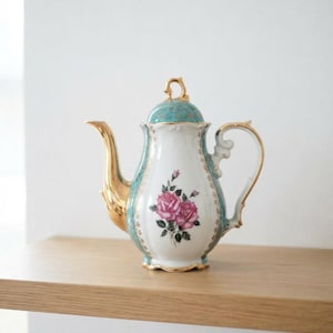Puede incluir: Una tetera de porcelana con cuerpo blanco y un motivo floral central de rosas rosas. La tetera tiene un cuello y una tapa de color cerceta, acentuados con bordes dorados en el asa, el pico, el remate de la tapa y la base con detalles dorados.