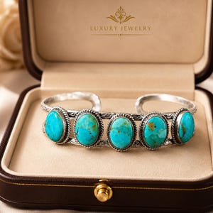May include: A silver cuff bracelet featuring five oval-shaped turquoise stones with brown matrix patterns. The metal band has a twisted rope border around each stone and engraved detailing along the sides. The text LUXURY JEWELRY is visible.