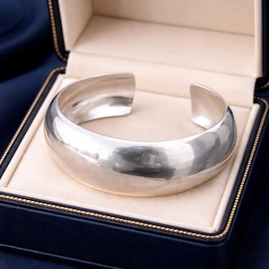 May include: A wide, polished silver cuff bracelet with a smooth, reflective surface and a rounded, open-ended design. The bracelet features a high-shine metallic finish and a substantial, curved band that maintains a consistent width throughout its form.