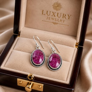May include: A pair of silver drop earrings featuring oval-shaped, faceted magenta-pink stones set in a twisted rope border. The earrings are displayed inside a dark brown wooden box with the gold text LUXURY JEWELRY printed on the interior lid.