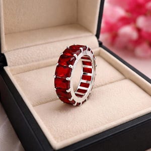 May include: A silver eternity band ring featuring a continuous row of rectangular-cut, deep red gemstones set in a prong setting. The ring is positioned upright inside a black jewelry box with a beige velvet interior lining.