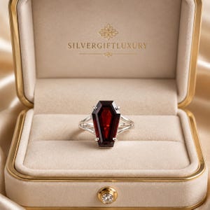 May include: A silver ring featuring a deep red, coffin-shaped gemstone held by four prongs, presented in a beige velvet-lined box with the text SILVERGIFTLUXURY printed in gold on the interior lid and a small circular crystal on the front latch.