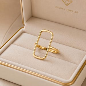 May include: A gold-toned ring featuring a prominent, vertically oriented rectangular open frame design. The ring is displayed inside a beige jewellery box with the text LUXURY JEWELRY printed in gold on the interior lid above a diamond icon.